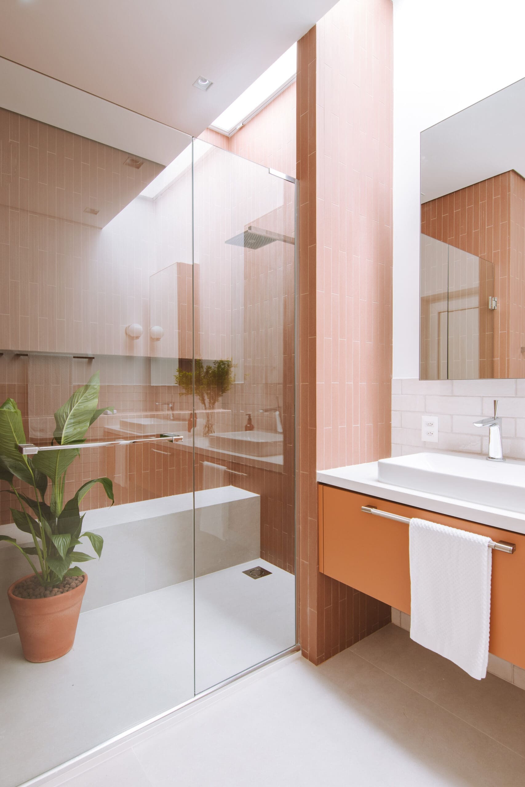 Banheiro moderno com box de vidro, revestimento em azulejos verticais terracota, pia com gabinete em cor terracota, espelho amplo e planta decorativa.