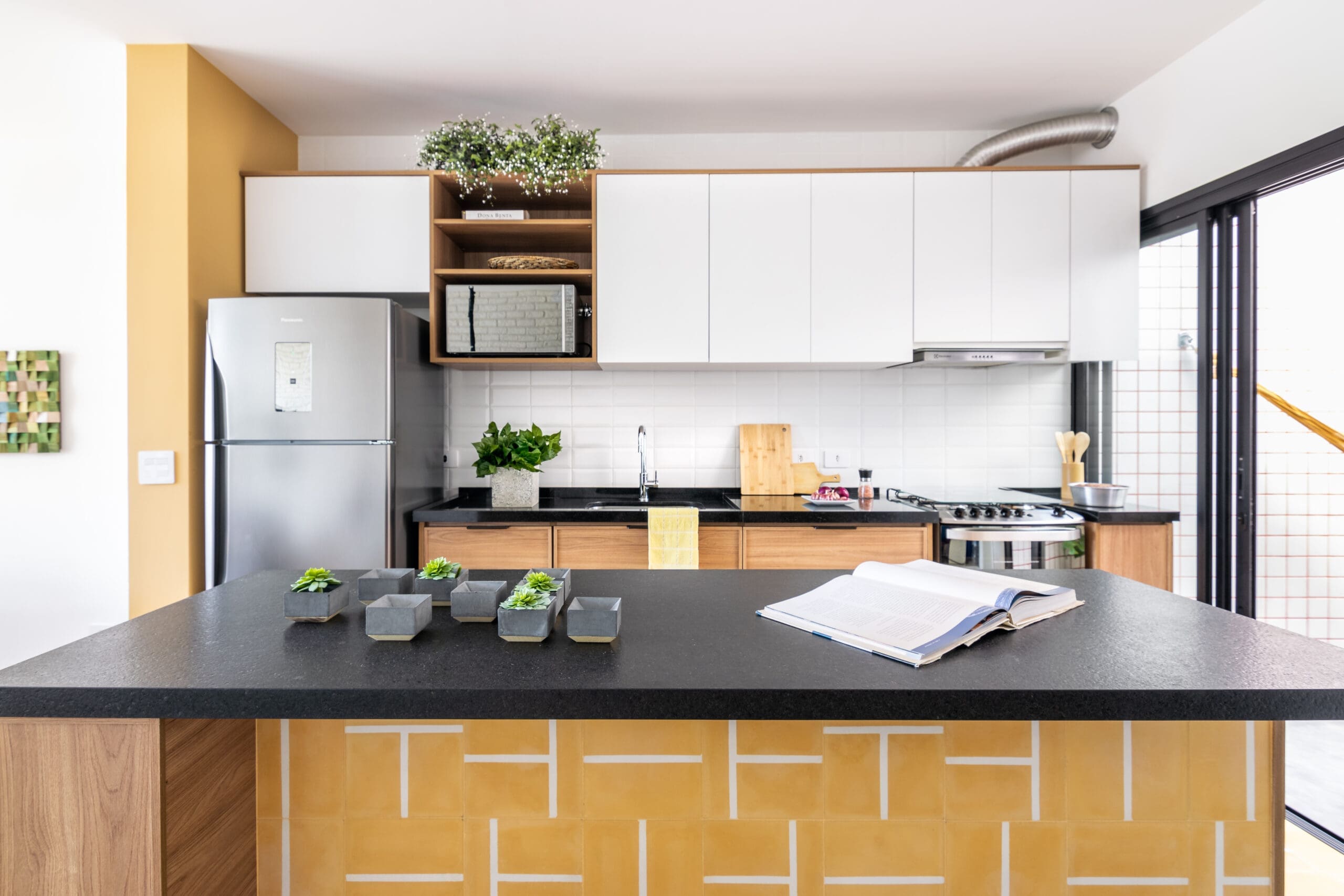 Cozinha moderna com bancada preta decorada com vasos de plantas e livro aberto, armários brancos e amadeirados, fogão, geladeira em aço inox e detalhes em ladrilhos amarelos.